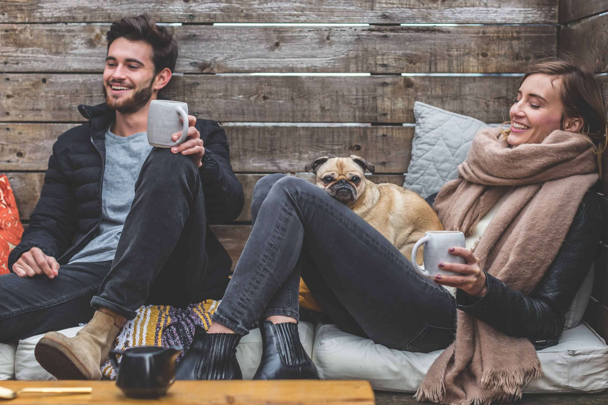 A man and a woman lounge on cushions with coffee and their pug